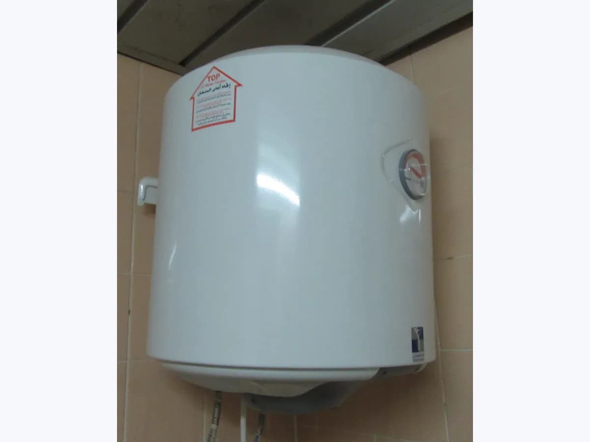 White cylindrical electric storage water heater mounted on a wall, with a front thermometer and water hoses below